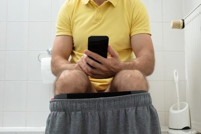 Moški sedi na straniščni školjki in ima v roki mobilini telefon.