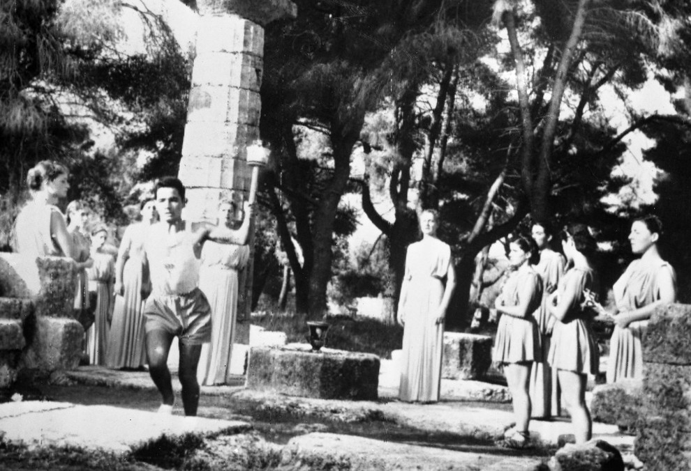 Olimpijski ogenj zapusti ravnice Olimpije. Dekleta, oblečena v starogrške noše, obkrožajo sveti oltar na ravnicah Olimpije, medtem ko prva svečenica …